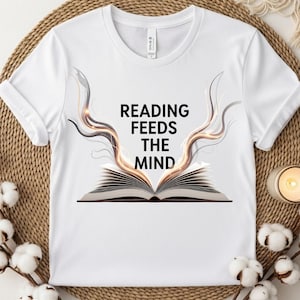 May include: A white short-sleeved t-shirt featuring a graphic of an open book with swirling, light-colored energy rising from the pages. The text "READING FEEDS THE MIND" is printed in black, bold, sans-serif capital letters above the book.