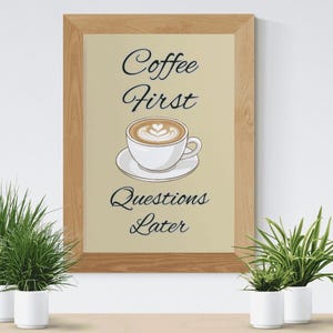 May include: A framed wall art print featuring a light beige background with a central illustration of a white coffee cup on a saucer. The text reads Coffee First above the cup and Questions Later below the cup in a black cursive script font.