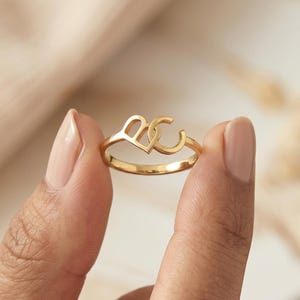 May include: A gold-toned ring featuring a polished metal band with a decorative centerpiece shaped like a stylized heart intertwined with the letter C. The design is crafted from a smooth, reflective material with a consistent metallic finish.