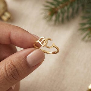 May include: A gold-toned ring featuring two interconnected capital letters, B and C, forming the centerpiece of the band. The metal has a polished, reflective finish, and the design consists of a thin, circular band that supports the letter motif.