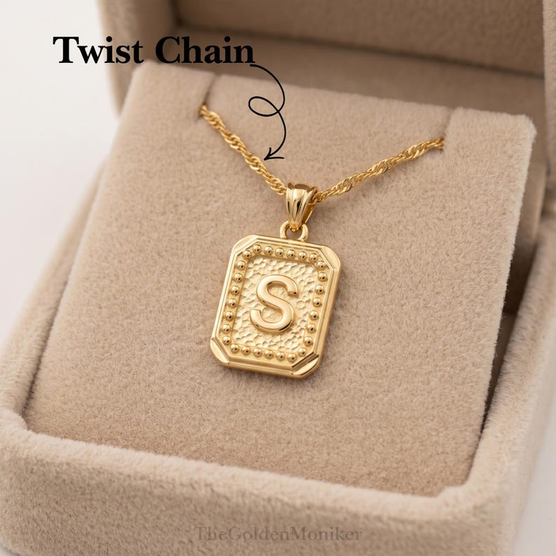 May include: Gold-colored necklace with a rectangular pendant featuring the letter "S". The pendant has a beaded border and is suspended from a twist chain. The text "Twist Chain" is above the necklace.