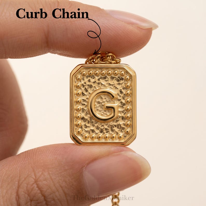 May include: Gold-colored rectangular pendant necklace with the letter "G" in the center. The pendant has a textured surface and is attached to a curb chain. The text "Curb Chain" is above the pendant.