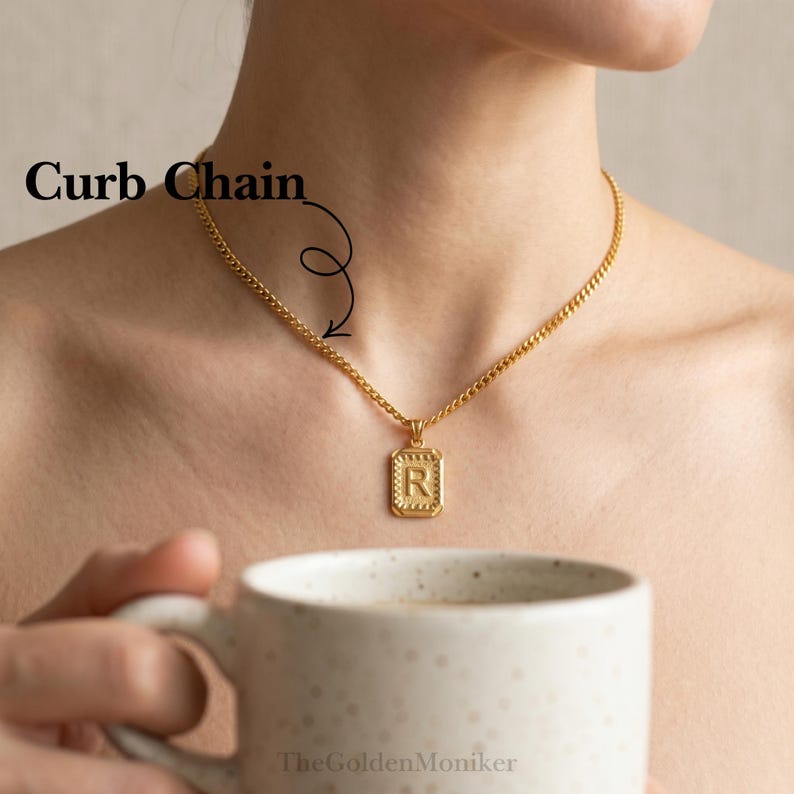 May include: Gold-colored curb chain necklace with a rectangular pendant featuring the letter "R". The pendant has a textured border. The necklace is worn around the neck.