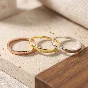 May include: Three thin metal rings in rose gold, yellow gold, and silver tones. Each ring features a smooth, polished band with a custom engraving on the outer surface. The rose gold ring is engraved with Maria, the yellow gold with Emma, and the silver with David.