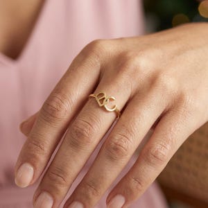 May include: A gold-toned ring featuring two interconnected letters, B and C, forming a central focal point on a thin, polished band. The design showcases a clean, metallic finish with the letters positioned side-by-side in a stylized, open font.