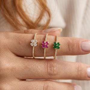 May include: Three gold rings, each featuring a four-leaf clover design. The first ring has clear white stones, the second has vibrant pink stones, and the third has deep green stones. Each thin band is encrusted with small, sparkling clear stones.