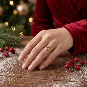 May include: A gold-toned ring featuring the letters B and C as the focal point of the band. The ring is worn on a finger, showcasing the metallic finish and the distinct shape of the two uppercase letters connected together in a simple design.