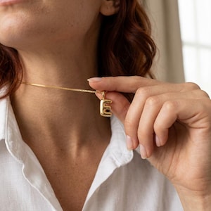 May include: A gold-toned necklace featuring a small, three-dimensional letter E pendant with a smooth, rounded surface. The pendant is suspended from a delicate, thin gold-toned box chain that rests against the neck.