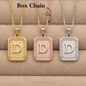 May include: Three initial pendant necklaces in gold, rose gold, and silver. Each necklace features a rectangular pendant with the letter "D" in the center, surrounded by a dotted border. The necklaces hang on box chains. The text "Box Chain" is visible.