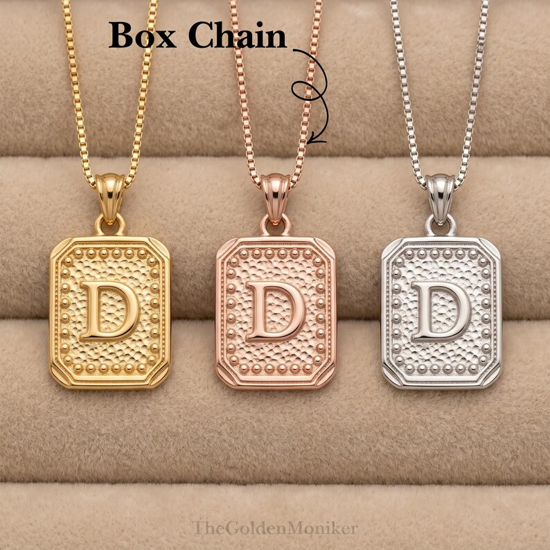May include: Three initial pendant necklaces in gold, rose gold, and silver. Each necklace features a rectangular pendant with the letter "D" in the center, surrounded by a dotted border. The necklaces hang on box chains. The text "Box Chain" is visible.