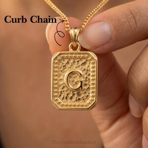 May include: A gold-colored pendant necklace with a curb chain. The rectangular pendant features the letter "G" in the center. The pendant has a textured surface and a beaded border. The text "TheGoldenMoniker" is at the bottom.