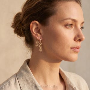 May include: Gold-colored dangle earrings featuring floral and geometric shapes. The earrings are attached to small hoop closures. The earrings are worn by a person with their hair up in a bun.