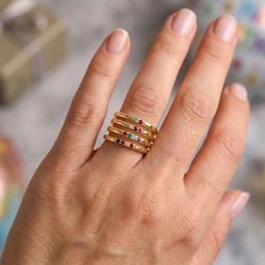 May include: A hand wearing five gold-colored rings. Each ring has a thin band and is adorned with small, colorful gemstones. The gemstones include blue, red, white, and green, adding a vibrant touch to the jewelry.