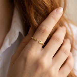 May include: A gold-toned open ring featuring two ends, each engraved with a single letter. One end displays the letter S, and the other end displays the letter Z. The band has a smooth, polished finish and is designed to be worn on a finger.