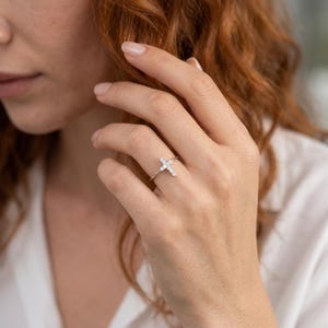 May include: A thin silver-toned ring featuring a small cross-shaped centerpiece adorned with multiple clear, sparkling stones. The cross is positioned horizontally across the band, which rests on a finger, showcasing a delicate and refined design.
