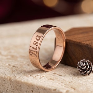 May include: A rose gold ring with the name "Lisa" engraved on the outside. The ring is displayed on a textured surface with a small pine cone and a wooden block in the background. The ring is a simple band design.