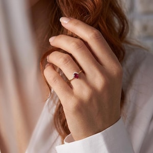 May include: A gold ring featuring a pear-shaped red gemstone as the centerpiece, accented by a small arc of clear white stones set along the top edge of the band. The thin, polished gold band holds the stones securely in a delicate, refined arrangement.