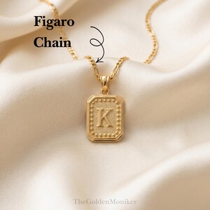 May include: A gold-colored Figaro chain necklace with a rectangular pendant. The pendant features the letter "K" in the center, surrounded by a dotted border. The necklace rests on a cream-colored satin fabric.