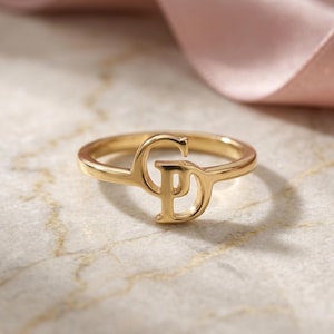 May include: A gold-toned ring featuring a polished metal band with two interconnected letters, C and D, forming a central monogram design. The letters are integrated into the circular band, creating a continuous, smooth, and reflective metallic surface.