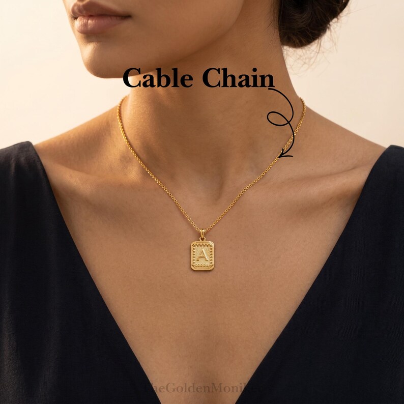 May include: A gold-colored cable chain necklace with a rectangular pendant. The pendant features the letter "A" in a decorative font. The necklace is worn on a person with a dark top. The text "Cable Chain" is visible.