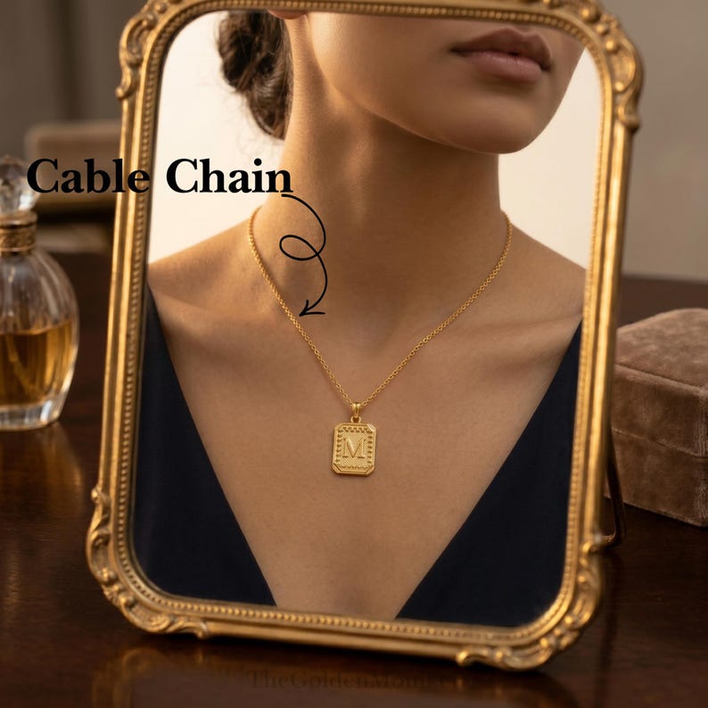 May include: A gold-colored cable chain necklace with a rectangular pendant featuring the letter "M". The necklace is reflected in a gold-framed mirror. The text "Cable Chain" is above the necklace.