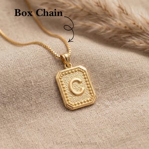 May include: Gold-colored box chain necklace with a rectangular pendant. The pendant features the letter "C" in the center, surrounded by a dotted border. The words "Box Chain" are at the top of the image.