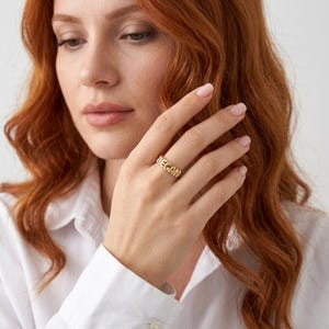 May include: A gold-toned ring featuring the name MEGAN in raised, block-style capital letters. The band is polished and metallic, encircling the finger with the personalized name clearly displayed across the front of the ring's surface.