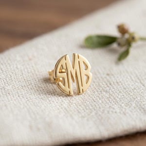 May include: A gold-toned circular stud earring featuring the cutout monogram letters S, M, and B. The letters are interconnected within a round frame, creating a polished metallic surface with a reflective finish and a post backing.