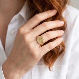 May include: A gold signet ring featuring a smooth, polished surface with an engraved monogram design on the circular face. The ring has a solid, tapered band that sits comfortably on the finger, showcasing a classic, metallic yellow gold finish.