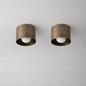 May include: Two cylindrical ceiling lights in an antique brass finish, each with a white bulb. The lights are mounted on a white ceiling. The design is minimalist and modern, suitable for various interior styles. The lights are approximately 15 cm in diameter.