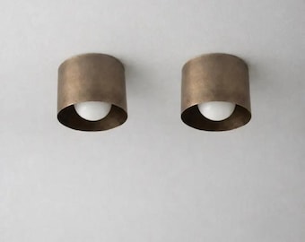 May include: Two cylindrical ceiling lights in an antique brass finish, each with a white bulb. The lights are mounted on a white ceiling. The design is minimalist and modern, suitable for various interior styles. The lights are approximately 15 cm in diameter.