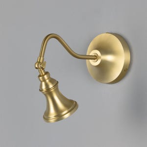 May include: A wall-mounted light fitting with a brushed brass finish. It features a circular wall plate, a curved arm, and an adjustable joint connecting to a bell-shaped lamp head. The design is metallic with a smooth, uniform golden tone.