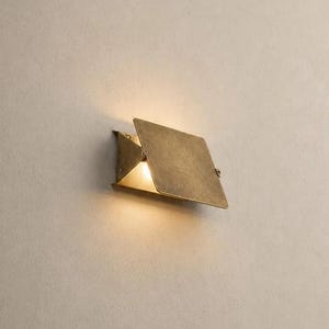 May include: A wall-mounted light fitting featuring a rectangular, brushed brass metal plate angled outwards. The fitting emits a warm, soft glow from behind the metal panel, casting light onto the wall surface above and below the central structure.