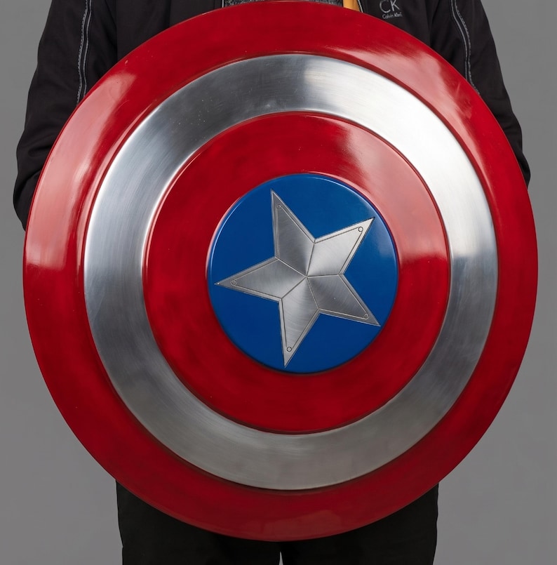 May include: A large, round shield in red, silver, and blue. The shield has a central blue circle with a silver star, surrounded by silver and red bands. A prop or collectible item.