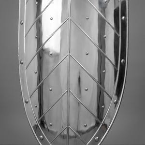 May include: A polished, silver-colored shield with a leaf-like design. The shield has a rounded top and bottom with a series of rivets around the edge. Diagonal lines and small rivets create a decorative pattern across the surface.