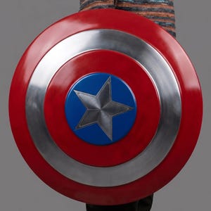 May include: A circular shield in red, silver, and blue. The shield has a central blue circle with a silver star. The outer ring is red, followed by a silver ring, and then another red ring. The shield is held by a person.
