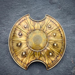 Könnte beinhalten: Ein goldfarbener Schild mit aufwendigen geprägten Mustern. Der Schild hat ein zentrales kreisförmiges Emblem mit detaillierten Mustern und ist mit Nieten und dekorativen Linien verziert. Das Design deutet auf ein historisches oder Fantasy-Thema hin.