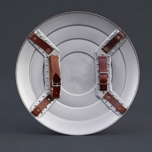 May include: A silver circular shield with concentric rings and brown leather straps. The shield has a metallic finish and is designed with riveted metal supports for the straps. The straps are secured with buckles.