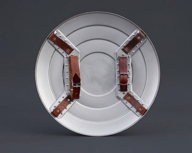 May include: A silver circular shield with concentric rings and brown leather straps. The shield has a metallic finish and is designed with riveted metal supports for the straps. The straps are secured with buckles.
