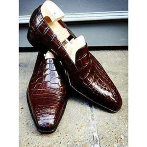 May include: A pair of dark brown loafers with a glossy, textured crocodile-skin pattern. Each shoe features a pointed toe and is fitted with a light-colored wooden shoe tree insert to maintain the shape of the footwear structure.