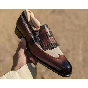 May include: A two-tone leather loafer featuring a dark brown burnished toe and heel, a beige suede vamp, and a decorative kiltie fringe panel. The shoe includes a dark brown leather strap with a silver-toned metal buckle across the instep.
