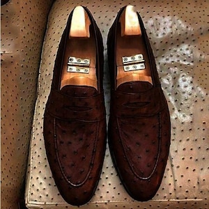 May include: A pair of dark brown loafers featuring a textured ostrich leather finish with a classic penny strap across the vamp. Each shoe contains a light-colored wooden shoe tree with a metal hinge mechanism inserted inside the interior.