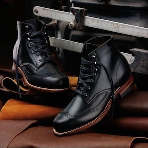 May include: A pair of black leather lace-up boots featuring a wingtip toe cap design, metal eyelets, and a stacked wooden heel with a visible welt. The boots have black laces and a smooth, polished finish, showcasing detailed stitching along the sides.