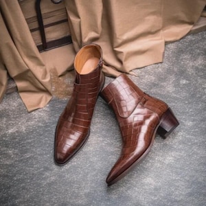 Könnte beinhalten: Ein Paar braune Stiefeletten mit glänzender Krokodilprägung, spitzer Zehenpartie und einem geschichteten Blockabsatz. Die Stiefel verfügen über einen seitlichen Reißverschluss und ein beiges Innenfutter, was eine strukturierte Silhouette ergibt.