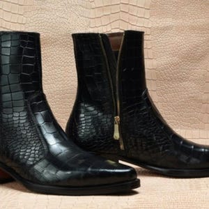 May include: A pair of black ankle boots featuring a glossy crocodile-embossed texture. The boots have a pointed toe, a stacked heel, and a gold-toned side zipper closure on the inner shaft. The design includes a smooth leather-like finish.