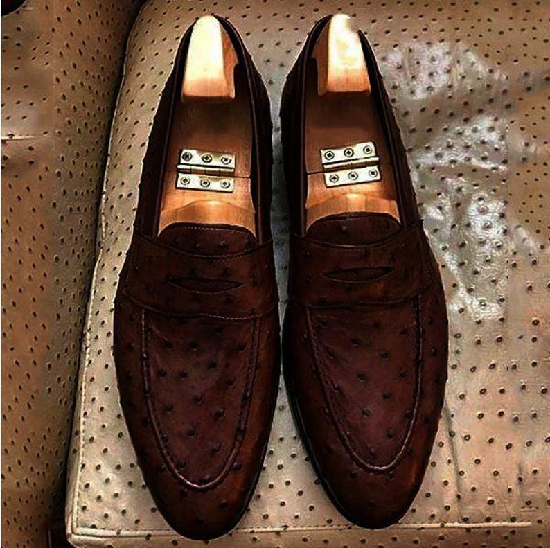 Peut inclure: Une paire de mocassins en cuir marron fonc&eacute; pr&eacute;sentant une texture &agrave; motifs de plumes sur toute la surface. Chaque chaussure comprend une bride horizontale sur l'empeigne et est &eacute;quip&eacute;e d'un embauchoir en bois clair avec charni&egrave;re.