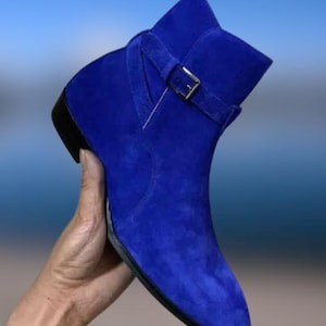Pode incluir: Uma bota de cano curto em camurça azul-real vibrante com biqueira pontiaguda, salto baixo preto e uma tira decorativa no tornozelo presa com uma fivela de metal prateado. O calçado apresenta uma textura uniforme e uma silhueta estruturada.