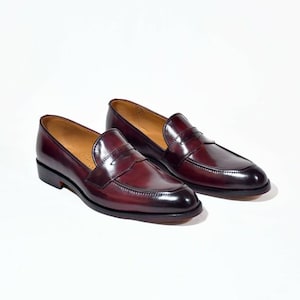 May include: A pair of deep burgundy leather penny loafers featuring a smooth, polished finish, a rounded toe, a classic strap across the vamp, and a low heel. The interior lining is tan, and the soles are dark with a visible stacked heel construction.