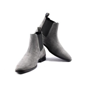 Könnte beinhalten: Ein Paar graue Chelsea-Boots aus Wildleder mit spitzer Zehenpartie, schwarzen elastischen Seiteneinsätzen und einer schwarzen Sohle. Die Stiefel verfügen über eine Zugschlaufe an der Rückseite des Knöchels und eine glatte, matte Oberfläche.