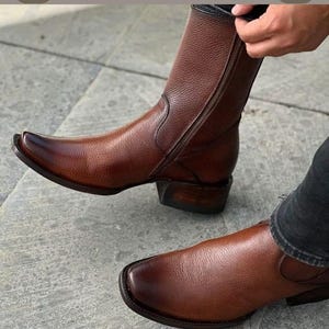 May include: A pair of brown leather boots with a textured grain finish. The boots feature a pointed toe, a stacked wooden heel, and a side zip closure. The leather has a polished, gradient-toned appearance, transitioning from a lighter brown to a darker, burnished shade at the toe and heel.